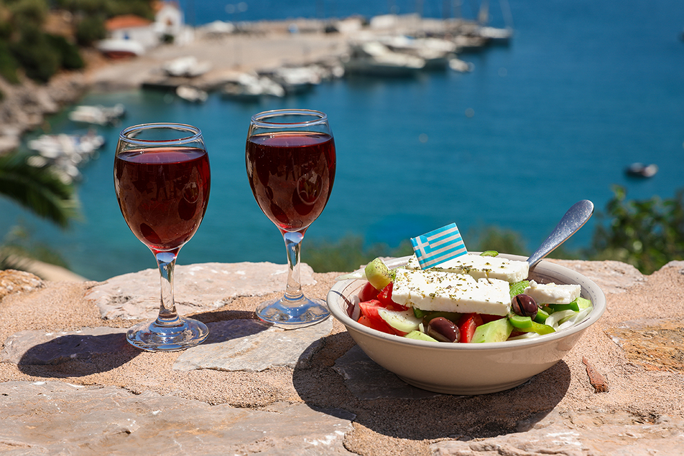 Two glasses of red wine and bowl of greek salad with greek flag on by the sea view, summer greek holidays concept.